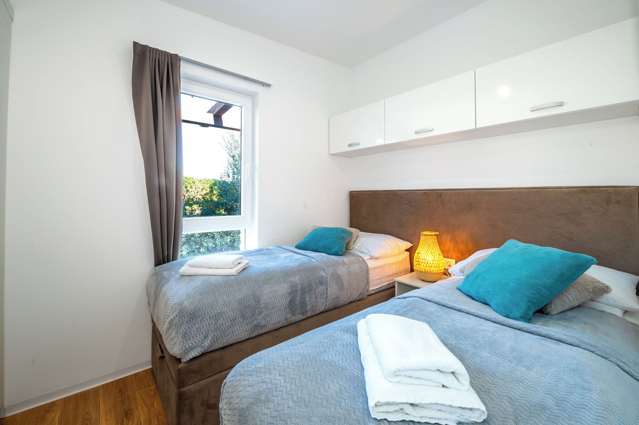 A double room with two beds, a window with curtains, a bedside table with a lamp, and white cabinets above the beds.
