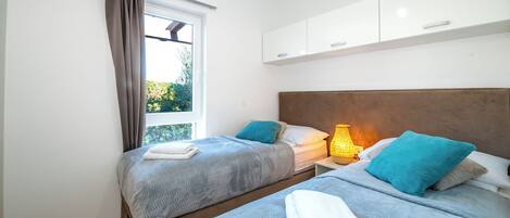 A double room with two beds, a window with curtains, a bedside table with a lamp, and white cabinets above the beds.