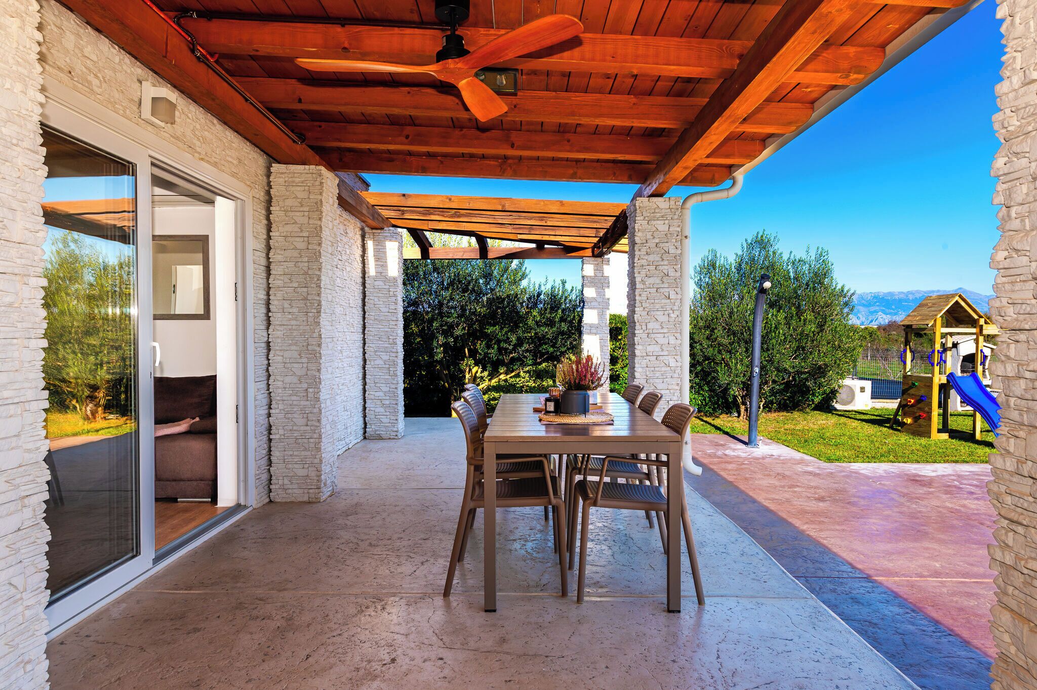 A covered patio with a dining table and chairs, a ceiling fan, and a view of a playground and mountains in the background.