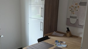 Eine kleine Küche mit Kühlschrank, einem Holzschrank, einem Esstisch mit zwei Platzdeckchen, einem schwarzen Stuhl und einer Wandvase mit Blumen.