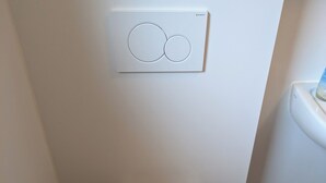 Ein kleines, modernes Badezimmer mit Toilette, Toilettenbürste und Wandspülknopf.