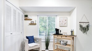 Una habitación con un armario blanco, un sofá color beige con una almohada azul, una mesita de madera con una cafetera, una planta en maceta y un cuadro en la pared.