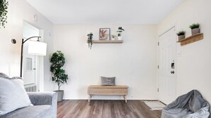 Una sala de estar moderna con un sofá gris, un banco y un sillón de peluche. Hay dos estantes con plantas y un cuadro en la pared.