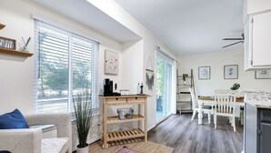 Una habitación luminosa y bien iluminada con una mesa de madera, sillas y una alfombra. Hay una ventana con persianas, una planta y un estante con un letrero.