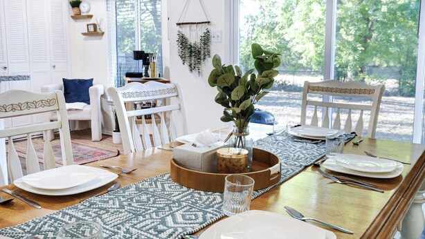 Un comedor con una mesa de madera dispuesta para cuatro, sillas blancas, un florero con follaje verde y vistas a árboles a través de amplios ventanales.