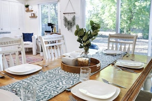 Un comedor con una mesa de madera dispuesta para cuatro, sillas blancas, un florero con follaje verde y vistas a árboles a través de amplios ventanales.