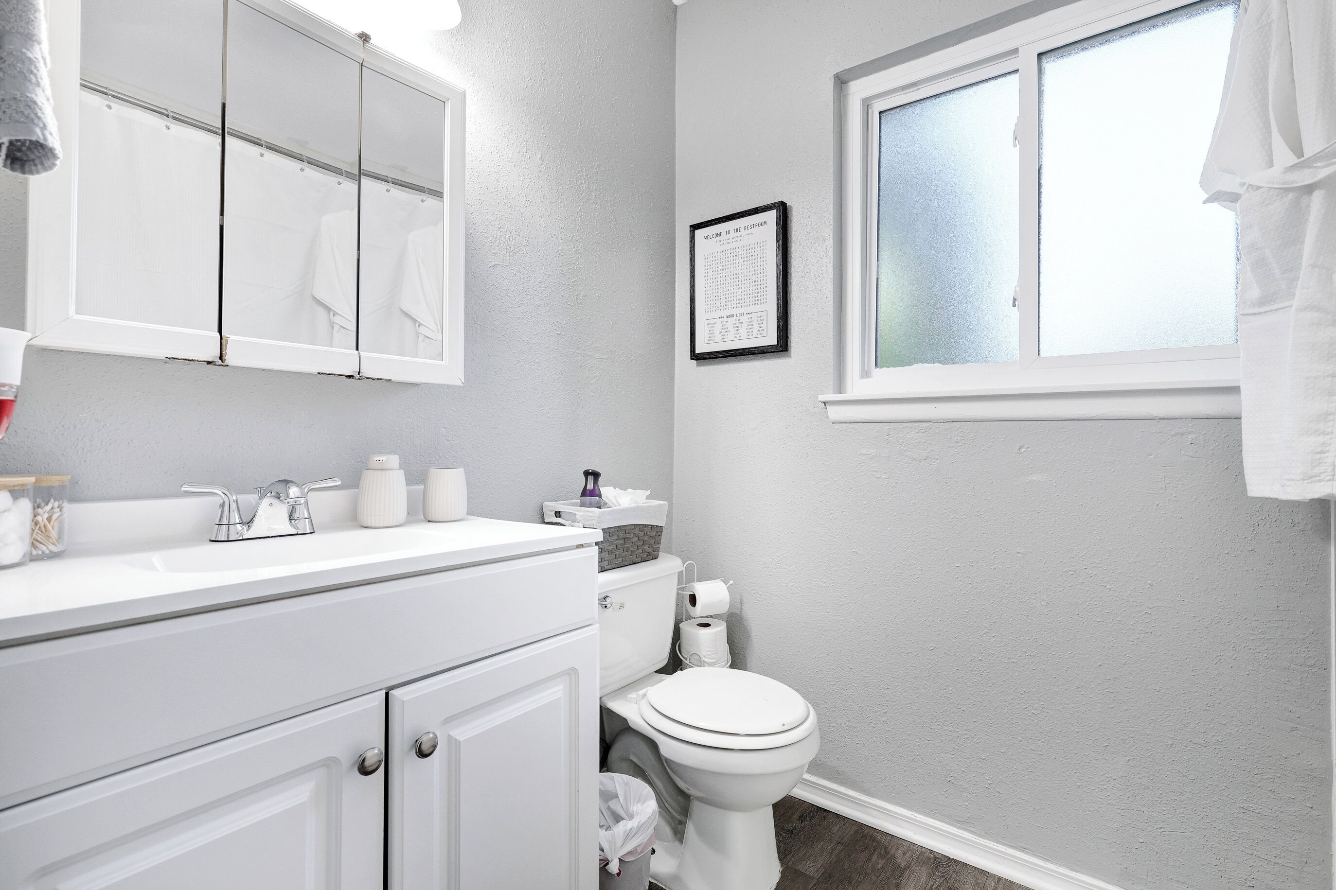 Ein Badezimmer mit weißem Waschbecken, Spiegel und Toilette. Ein Fenster mit Milchglas und ein gerahmtes Willkommensschild sind an der Wand angebracht.