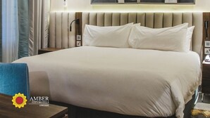 Quarto de hotel com uma cama grande, uma mesa de cabeceira, uma obra de arte na parede e uma janela com cortinas.