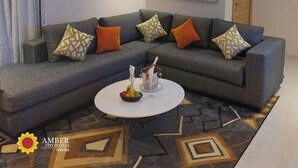 Uma sala de estar moderna com um sofá modular cinza, almofadas estampadas, uma mesa de centro branca com frutas e um copo, e uma grande pintura abstrata na parede.