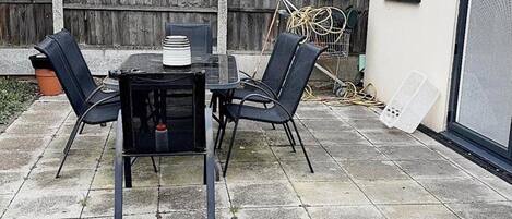 An outdoor patio with a table and four chairs, a wooden fence in the background, and a white wall with a window.