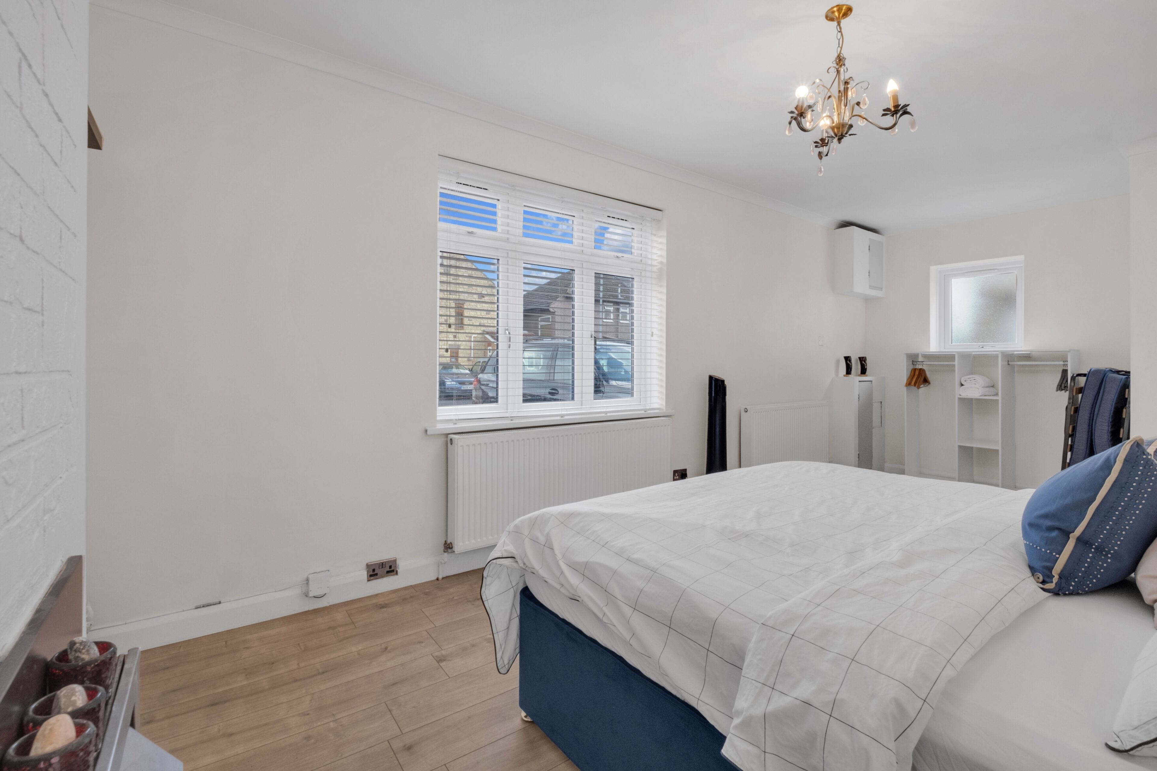 A bedroom with a large bed, a window with blinds, a radiator, a wardrobe, and a chandelier.