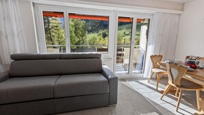 A modern living room with a grey sofa, a dining table with chairs, and a view of a green landscape through large windows. - Bright studio with mountain views (Leukerbad)
