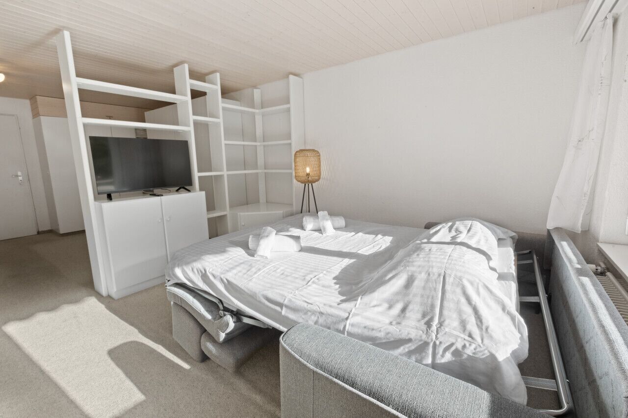 A modern, minimalist hotel room with a bed, a TV, a desk, and a lamp.