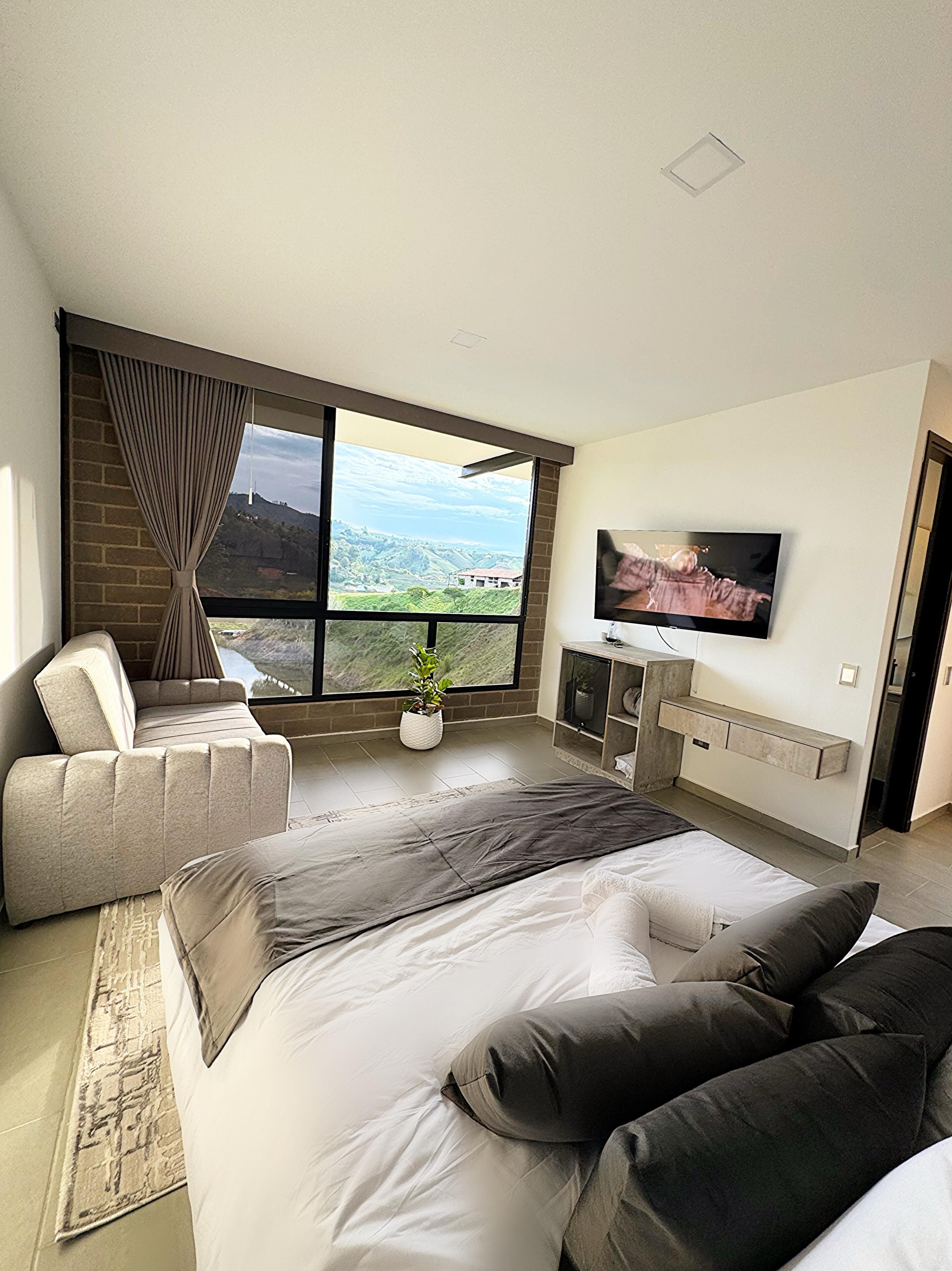 Une chambre moderne avec un grand lit, une télévision, un canapé et une vue sur un paysage montagneux, visible à travers une grande fenêtre.