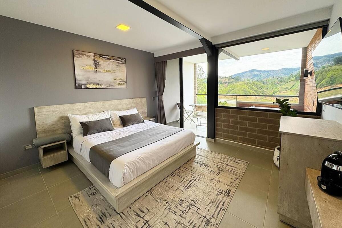 A modern bedroom with a large bed, a small bedside table, a nightstand, a view of the outdoors, and a balcony with a table and chairs.