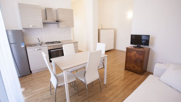 A compact kitchen with a refrigerator, stove, and cabinets. There is a dining table with chairs and a small TV on a wooden cabinet. - Anna Apartment just a few minutes from the centre (Portoferraio)