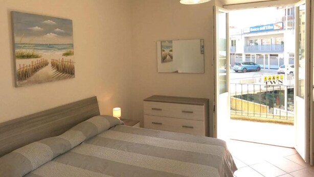 Ein Schlafzimmer mit einem Bett, einem Nachttisch, einer Lampe, einem Strandbild, einem Spiegel und Blick auf eine Straße und Häuser durch eine geöffnete Tür.