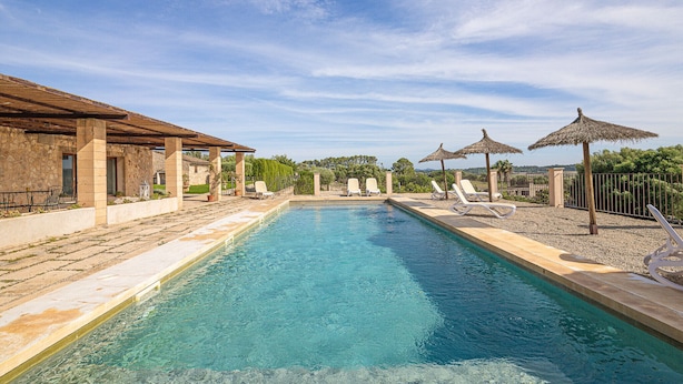 Une piscine rectangulaire à l’eau d’un bleu limpide, entourée de chaises longues et de parasols, avec en arrière-plan des bâtiments en pierre et des arbres sous un ciel dégagé.