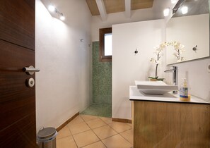 Une salle de bain avec une porte en bois, un lavabo blanc avec un miroir, une douche carrelée de carreaux verts et une petite fenêtre.