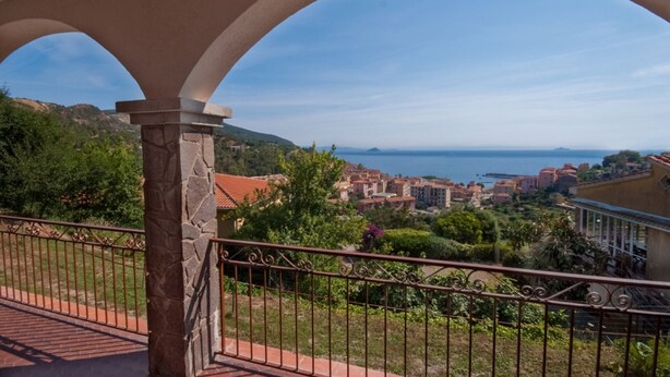 A balcony with stone pillars and wrought iron railing overlooking a coastal town with buildings and greenery. - Four-room apartment  with a view over Rio Marina (Rio Marina)
