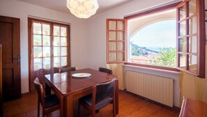 A dining area with a table and chairs, a hanging light fixture, and a window with an open shutter. - Four-room apartment  with a view over Rio Marina (Rio Marina)