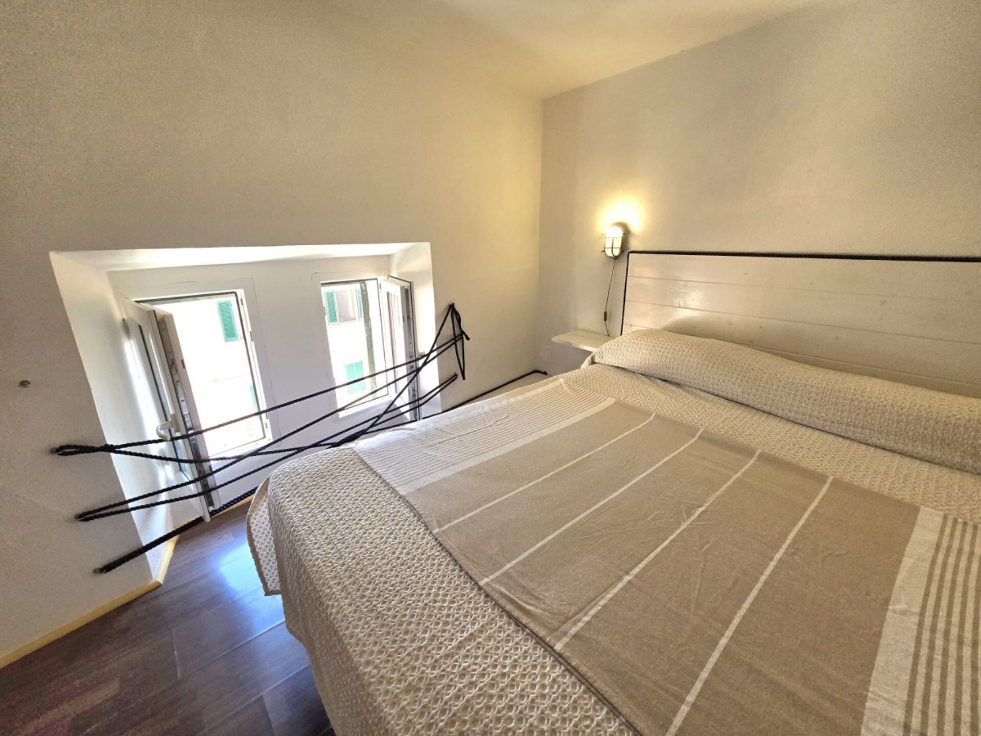 Ein Schlafzimmer mit einem Bett, einem Nachttisch, einer Wandlampe und Blick ins Freie durch ein Fenster.