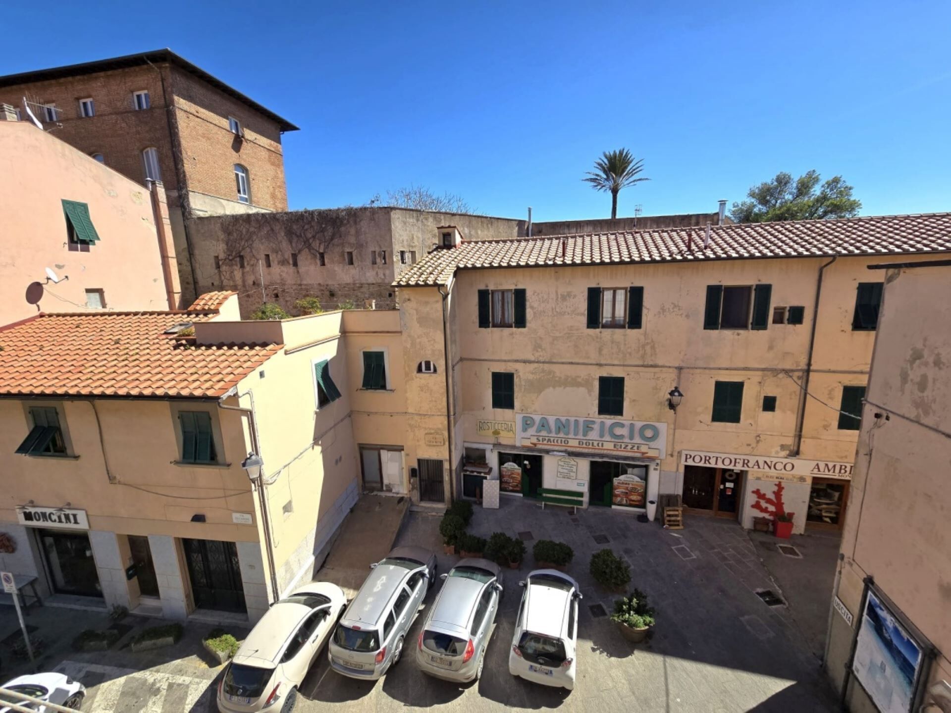 Vista di una strada con un edificio con la scritta "PANIFICIO" e "PORTOFRANCO AMBIENTE", auto parcheggiate e un cielo azzurro intenso.