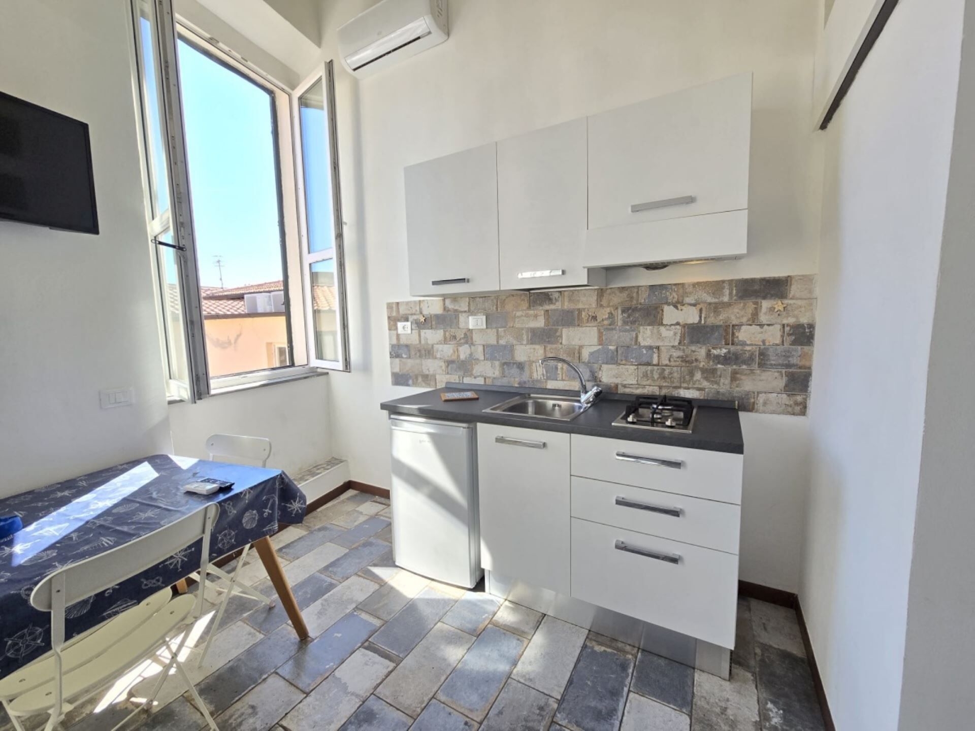Una cucina compatta con pensili bianchi, zoccolo in piastrelle e una finestra che lascia entrare luce naturale.