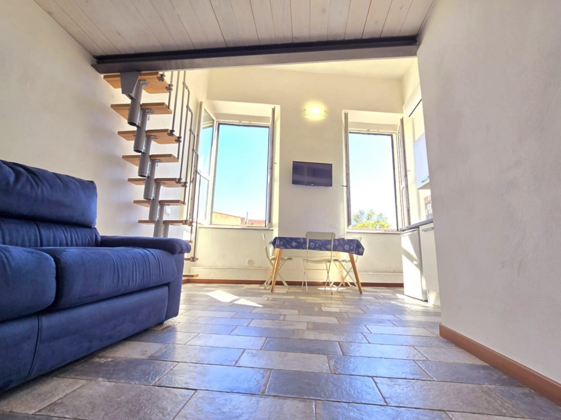 Ein Zimmer mit einem blauen Sofa, einem kleinen Tisch mit gemustertem Tischläufer und einer Treppe, die zu einer oberen Ebene führt.