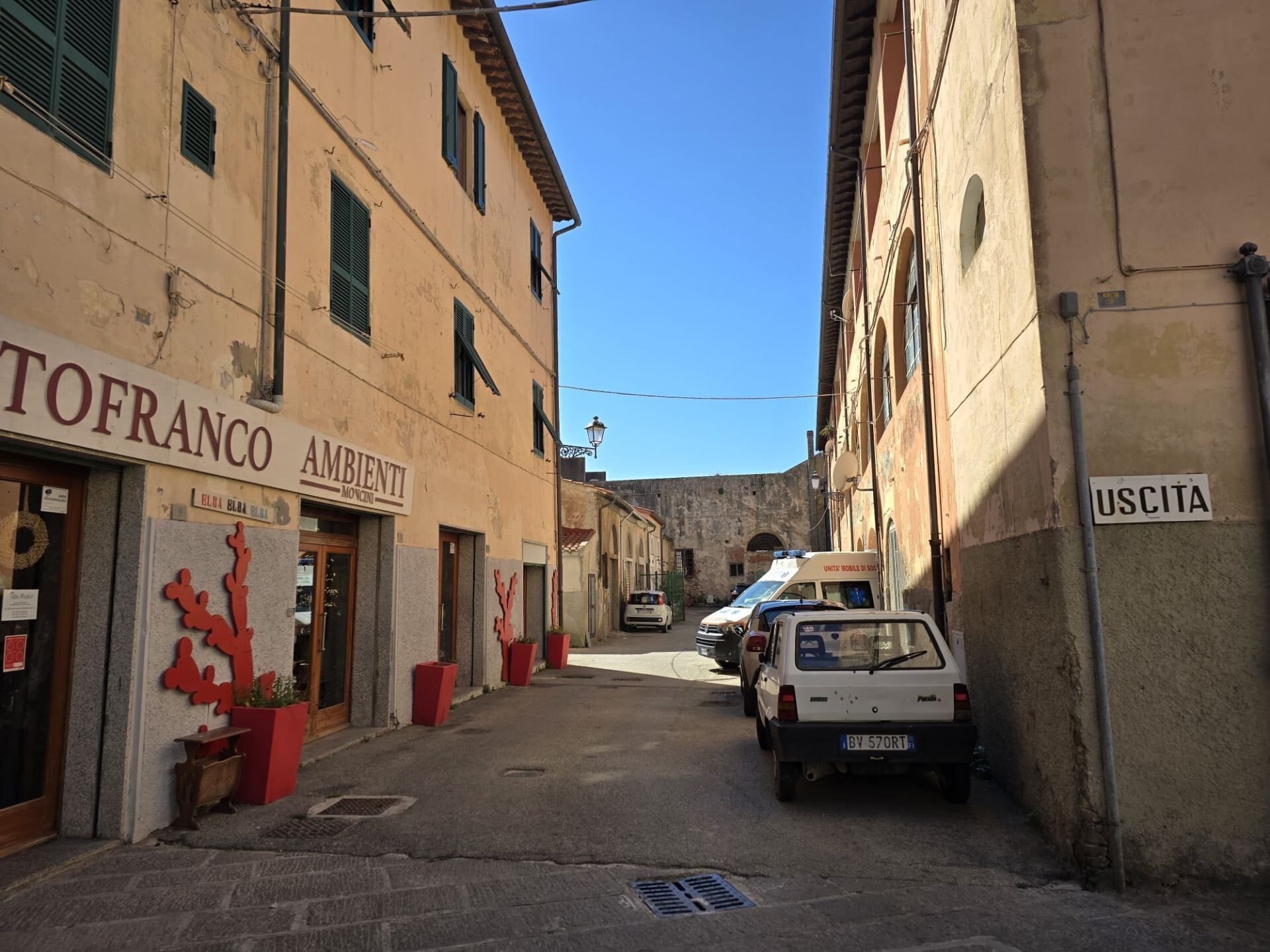 A narrow street with buildings on either side, including a shop named "TOFRANCO AMBIENTI" and a sign for "USCITA".