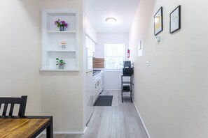 A narrow hallway leading to a kitchen with white cabinets, a dining table, and a small shelf with a plant.