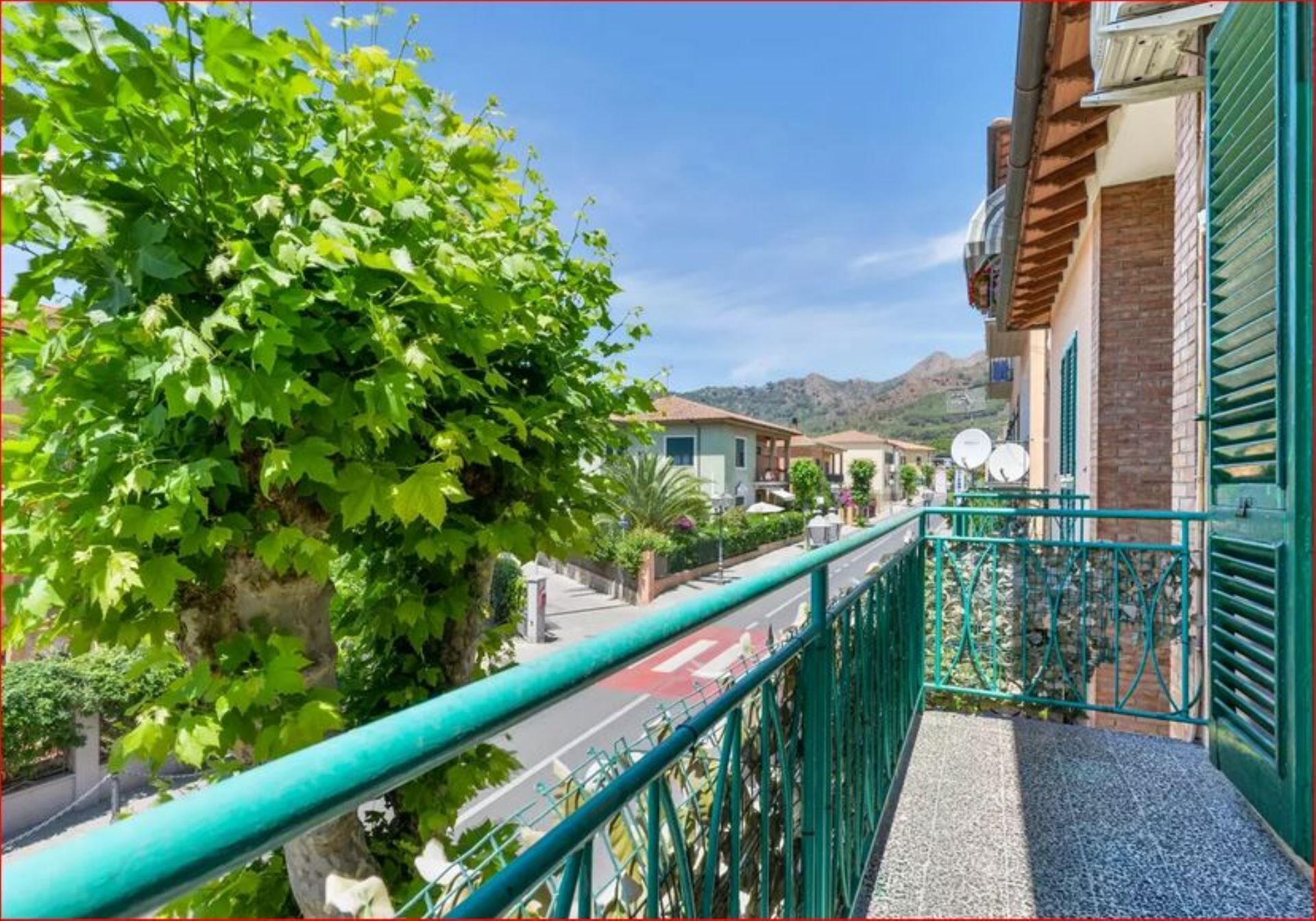 A balcony with a view of a street, a building with a tiled roof, and a mountain in the background.
