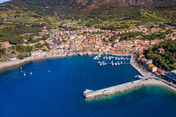 An aerial view of a coastal town with a marina, residential buildings, and hilly terrain in the background.
