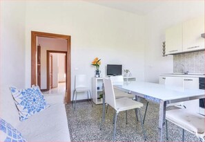 Un salon moderne et minimaliste avec un canapé blanc, une table à manger et des chaises. On y trouve également un téléviseur et une petite plante posée sur une étagère.