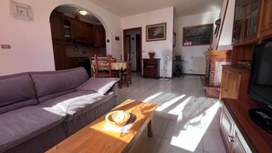 Un salon avec un canapé, une table basse en bois et un téléviseur. On y trouve également un coin repas avec une table et des chaises, et une cuisine à l’arrière-plan.
