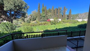 Un balcon doté d’un sol carrelé, d’une rambarde en métal et offrant une vue sur un jardin aménagé, avec différents arbres et arbustes.