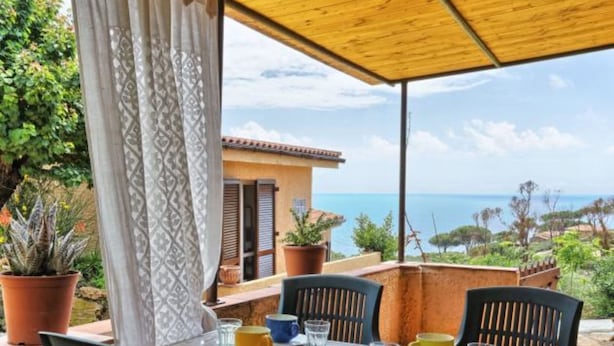 A table set for two with chairs, a wooden pergola overhead, and a view of the sea.
