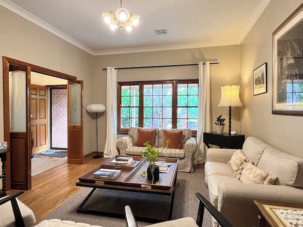A living room with a sofa, armchair, coffee table, and a potted plant. There is a window with curtains, a lamp, and a framed picture on the wall.