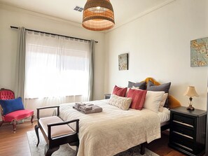 A bedroom with a bed, a chair, a nightstand, a lamp, and a window with curtains.