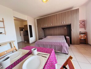 Un dormitorio compacto con cama, una mesita y una silla, un armario y una kitchenette al fondo.
