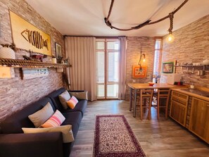 Een gezellige woonkamer met een donkere bank, een kamerbreed tapijt en een keukenhoek met houten kastjes en een eettafel.