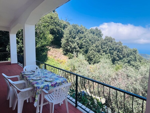 Un balcón con una mesa para dos, rodeado de sillas de plástico blanco, con vistas a un paisaje montañoso de exuberante vegetación.