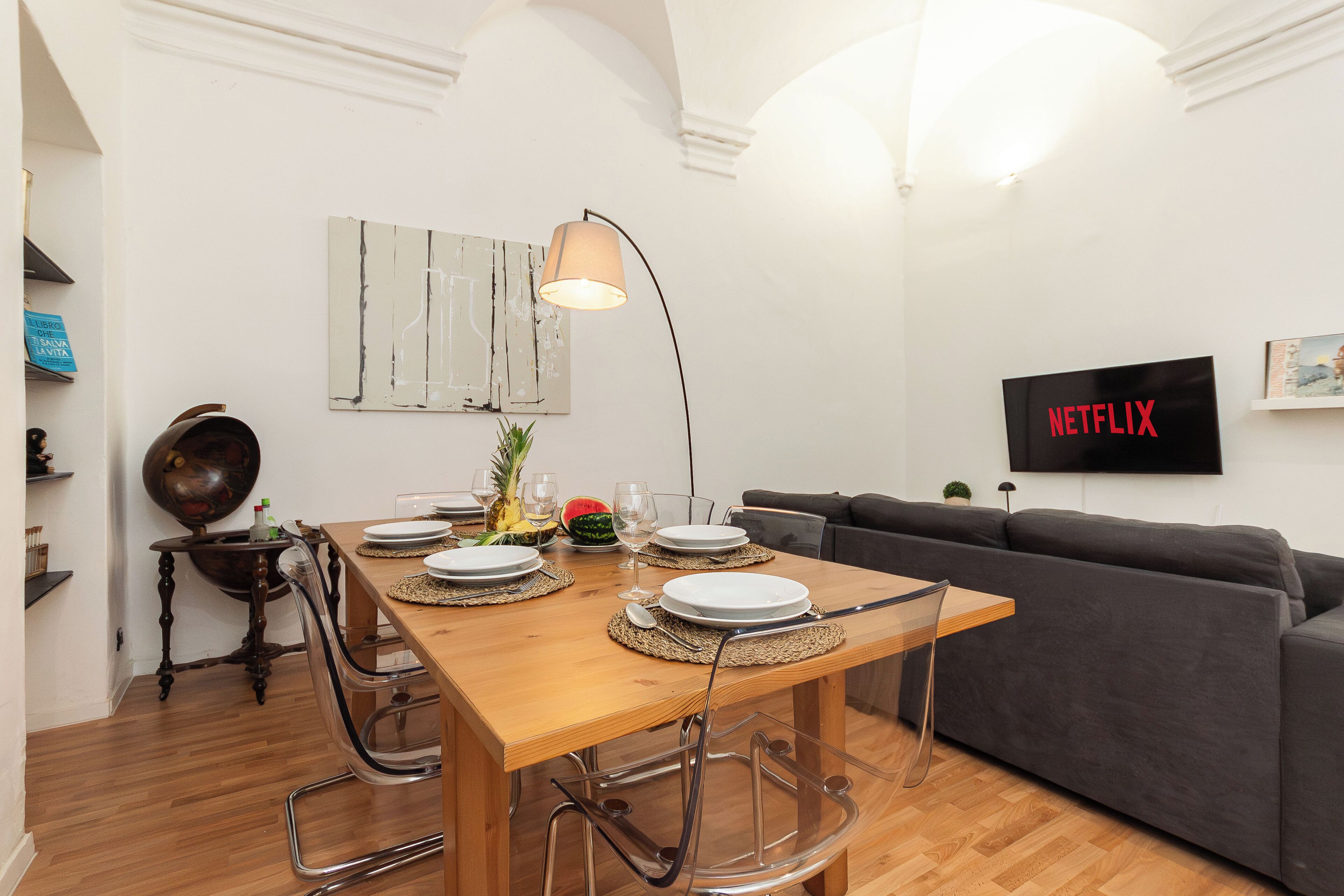 A dining area with a wooden table set for four, a dark grey sofa, and a flat-screen TV mounted on the wall.