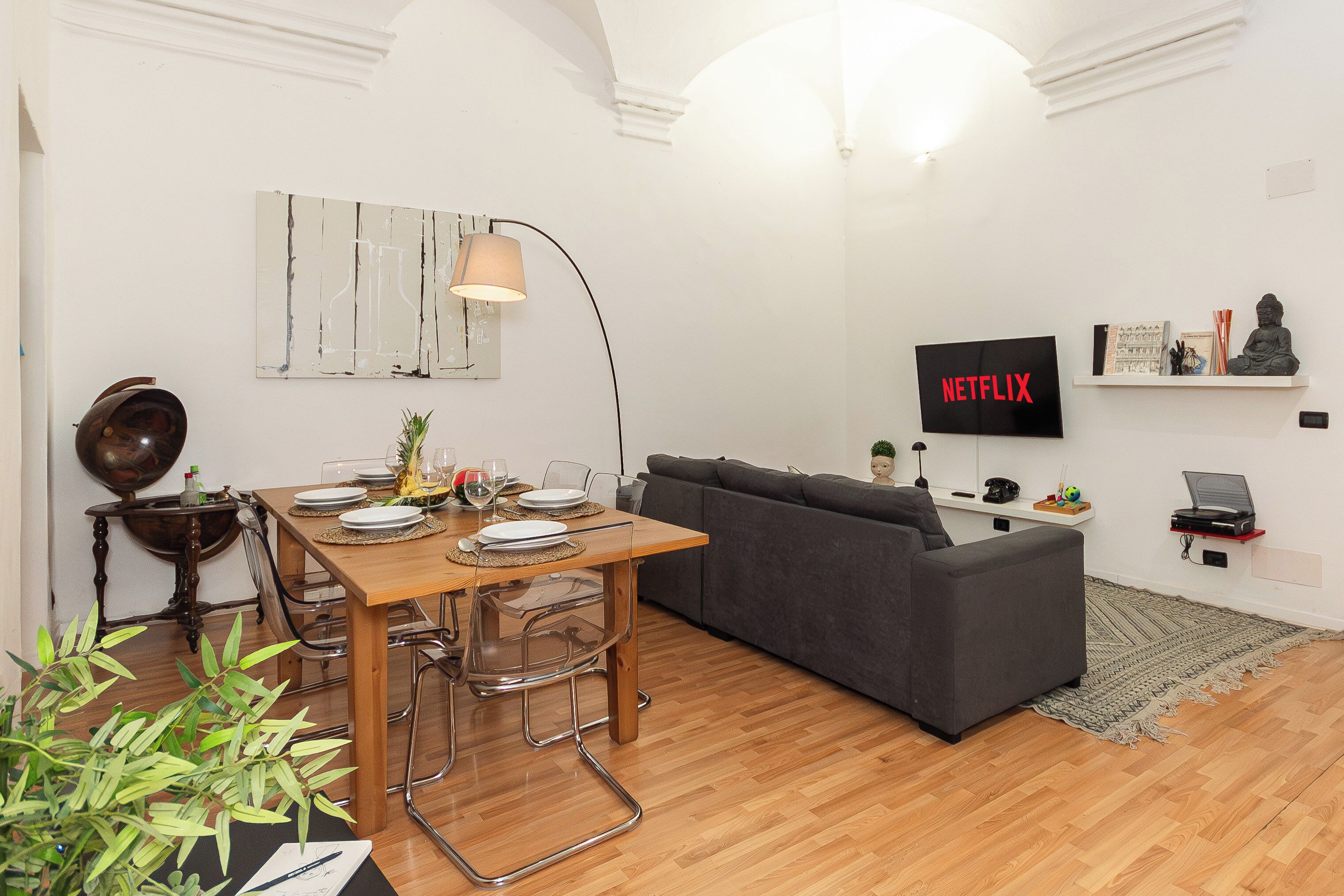 Un salon moderne comprenant un coin repas, un canapé gris, une table à manger en bois avec des chaises, une lampe et un téléviseur affichant le logo « NETFLIX ».