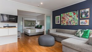Una habitación de hotel moderna con una cama grande, un sofá gris, un otoman gris y un televisor de pantalla plana sobre un mueble blanco.