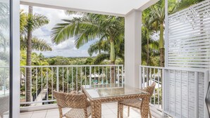 Un balcón con una mesa de vidrio y sillas de mimbre, con vista a un jardín exuberante con palmeras y una piscina.