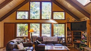 Ein gemütliches Wohnzimmer mit hoher Decke, großen Fenstern, einem Kamin und einer Bücherwand.