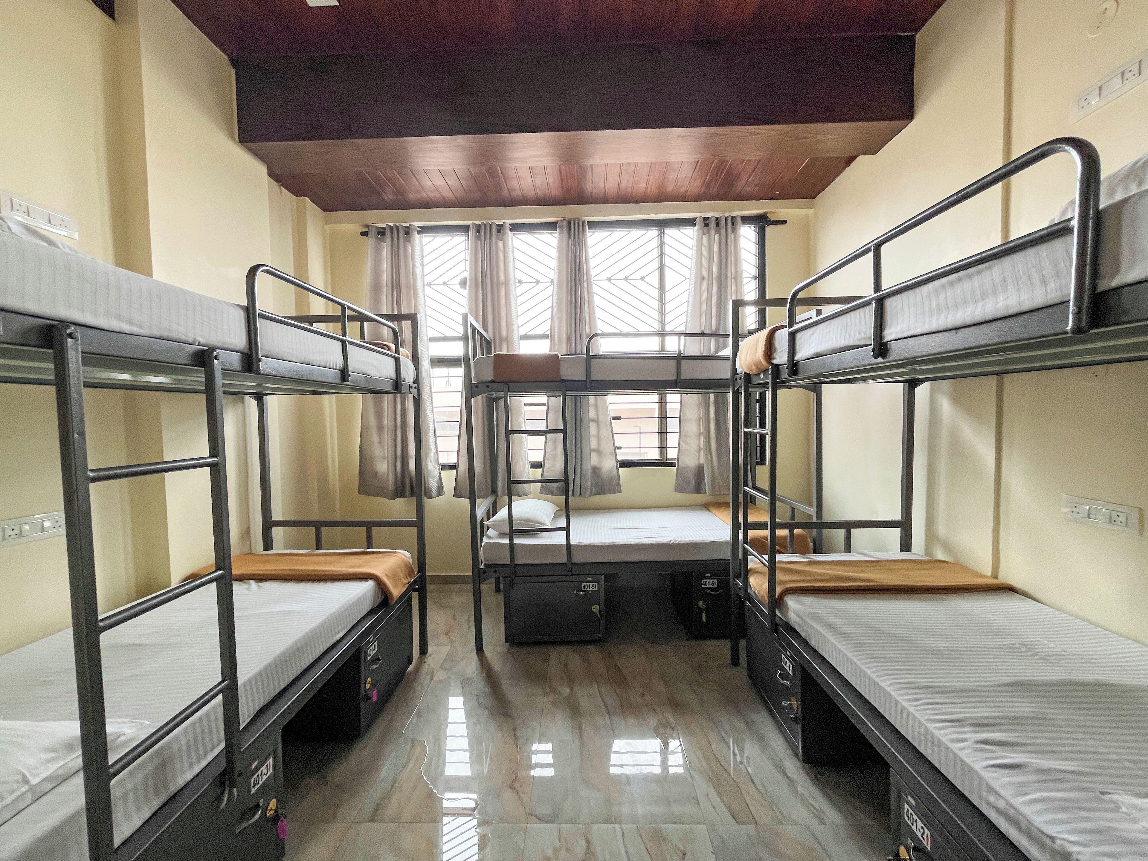 A dormitory room with bunk beds, a wooden ceiling, and a window with curtains.