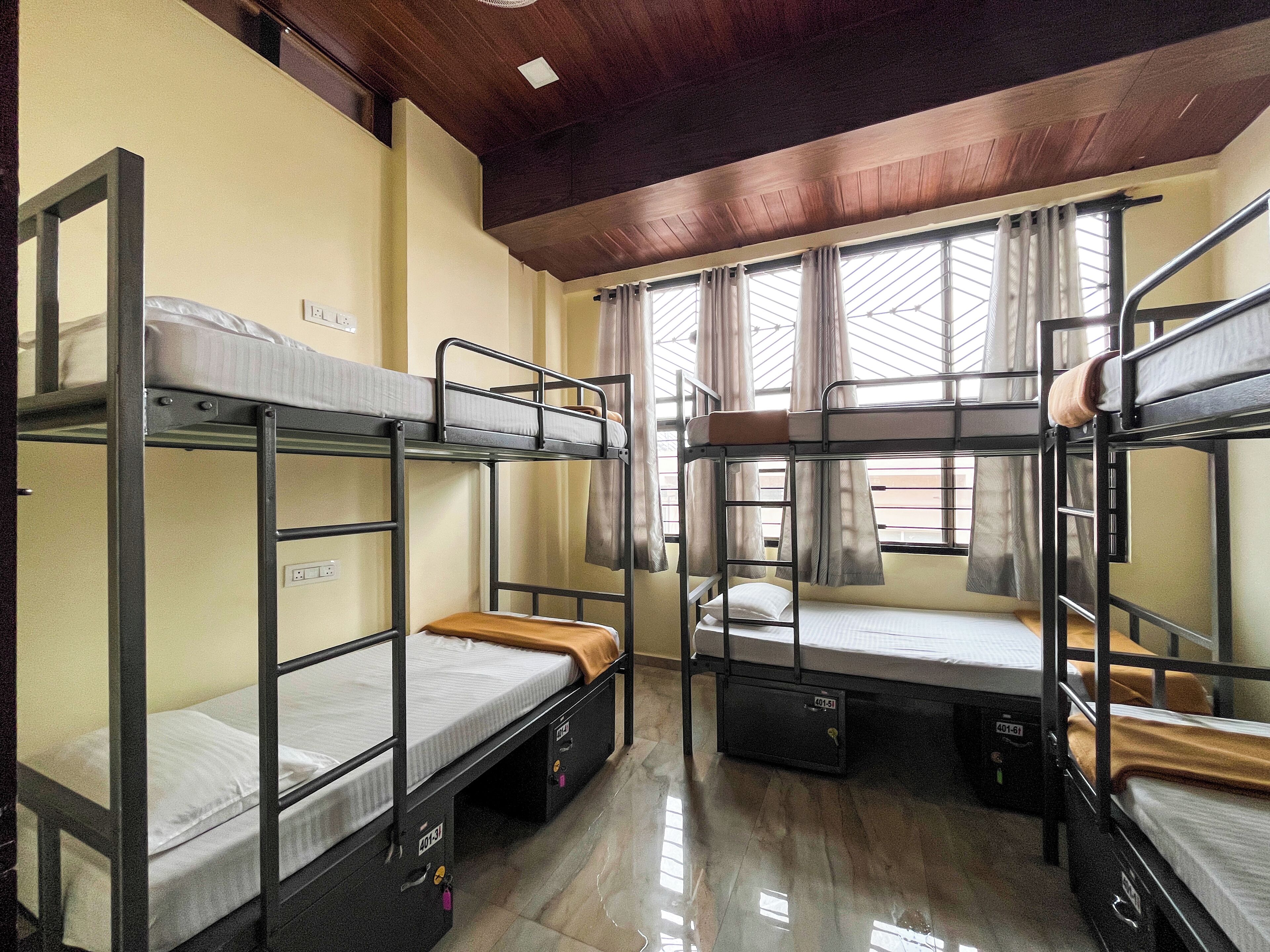 A dormitory room with bunk beds, a wooden ceiling, and a window with curtains.