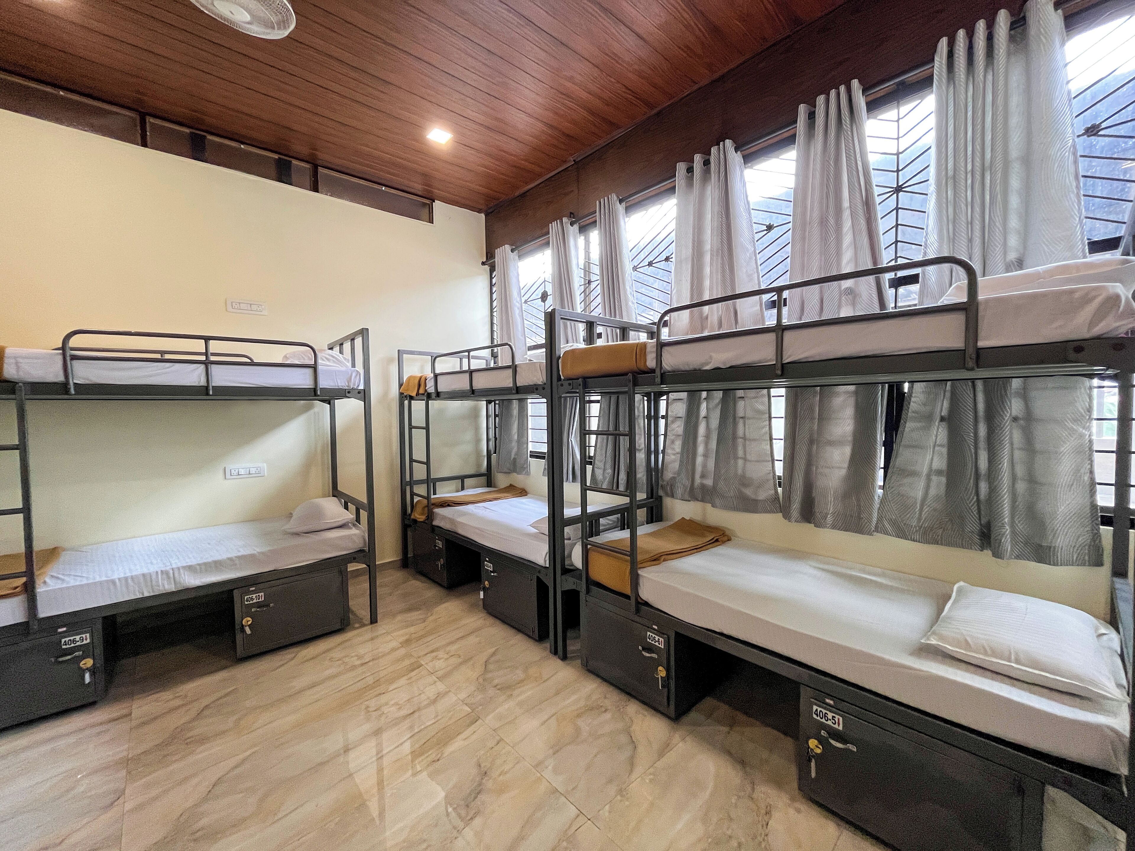 A dormitory room with bunk beds, wooden ceiling, and a window with curtains.